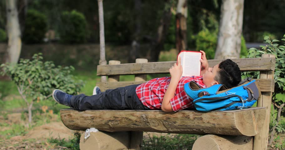 Portrait of a happy teen student with backpack sitting on a park bench and reading book in the summer green garden