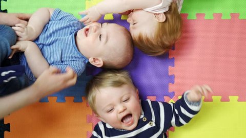 Overhead View Of Babies Lying On Mat At Nursery PlaygroupBeing Tickled By Mothers And Laughing