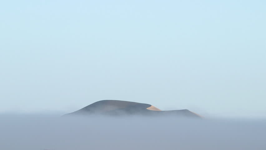 Tall sand dune in Namib Desert enveloped by fog with peaks rising out as fog gently moves across