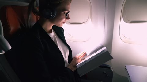 Close up happy smiling woman in digital headphones for noise cancellation enjoying flight reading interesting educative literature book while sitting near airplane window.Female passenger in aircraft