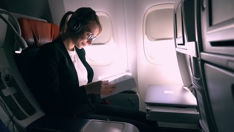 Positive young woman with modern headphones for noise cancellation listening audio music record and reading motivation book during flight time in airplane. Female aircraft passenger feeling carefree