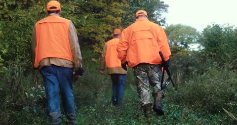 Three men in camouflage and blaze orange carrying rifles while hunting in the bush.