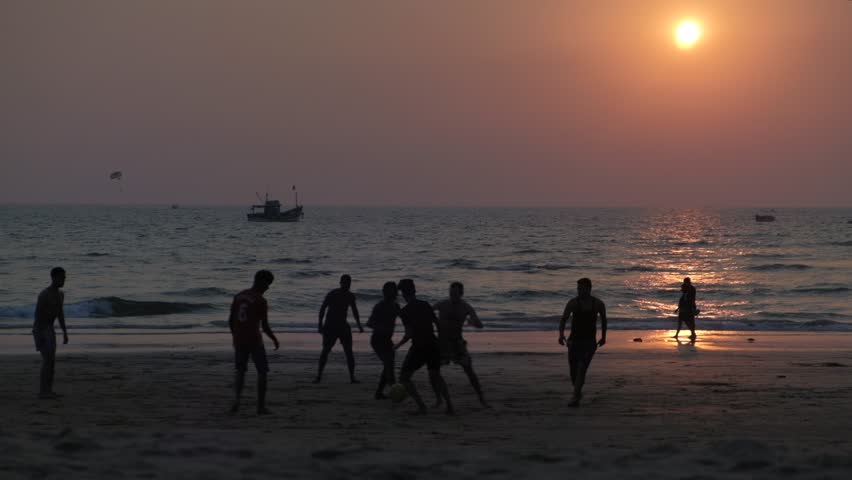 Silhouette of guys playing football on the beach while the sun is setting behind them in the Arabian sea with a ship and some small boats as seen from a beach in Goa, India 