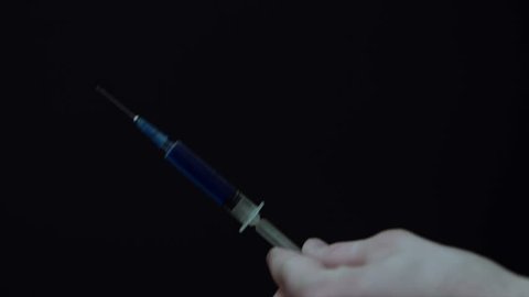 Closeup of a young man tapping the syringe with a blue liquid inside, then he sprinkles some of the liquid into the air. Isolated on black. Drug addict concept.