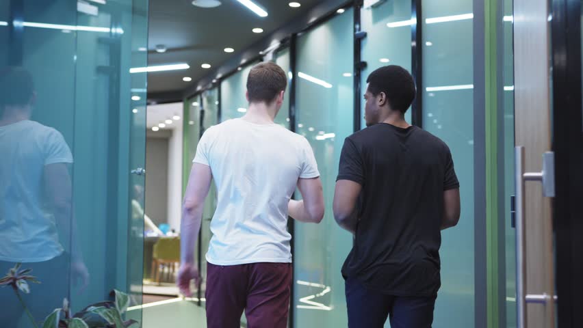 Rear view follow shot of two diverse office workers walking along corridor and talking on the move, greeting their female colleague leaving office and passing by manager using coffee machine Royalty-Free Stock Footage #1027227635