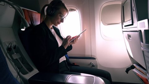 Caucasian female passenger in digital bluetooth headphones for noise cancellation chatting online via digital tablet using wifi internet connection on board. Young woman browsing network during flight
