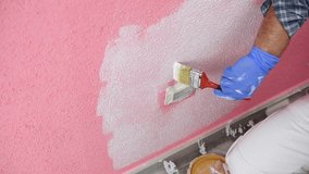 Caucasian house painter worker in white overalls, paint the pink wall with the white paint with the small brush. Construction industry. Footage.