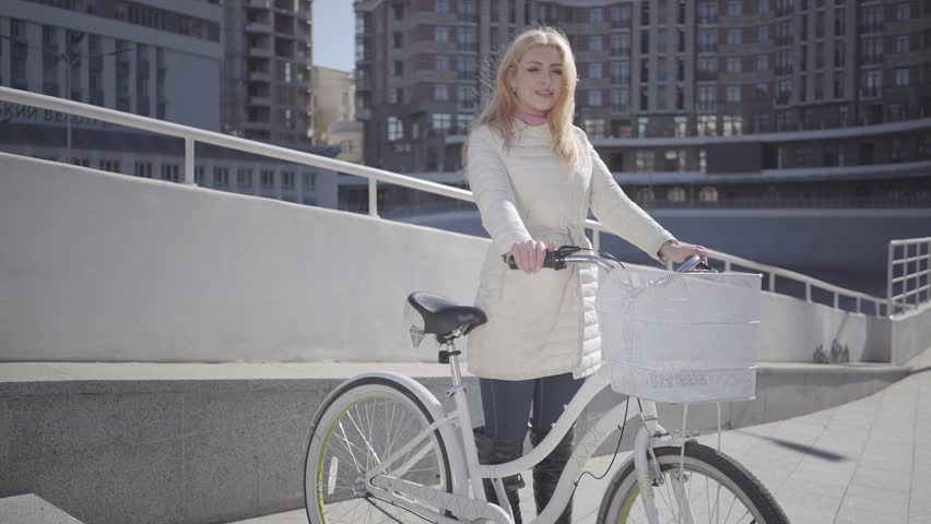 Pretty blond woman in warm white jacket standing at the city street with bicycle looking in the camera touching her hair. Leisure of young city dweller. Active lifestyle. Slow motion