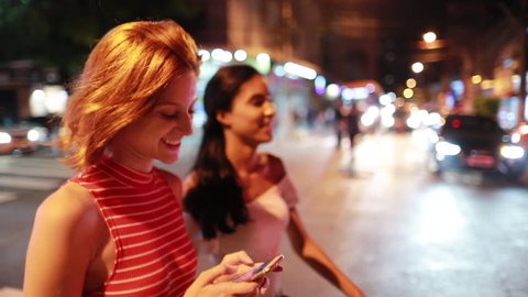 
Two multiracial female friends walking at night, young woman shows cellphone screen to friend, crossing street smiling