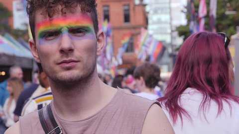 Guy with serious eyes and facepaint on at LGBTQ Pride, Lesbian, Gay, Bisexual, Transgendered, Queer, Parade with rainbow flags and slow motion
