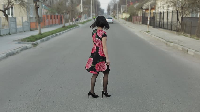 Attractive young woman in a dress with flowers turns to camera and smiles seductively, continues walking away from the camera. Cute girl walking on the highway. Slow motion