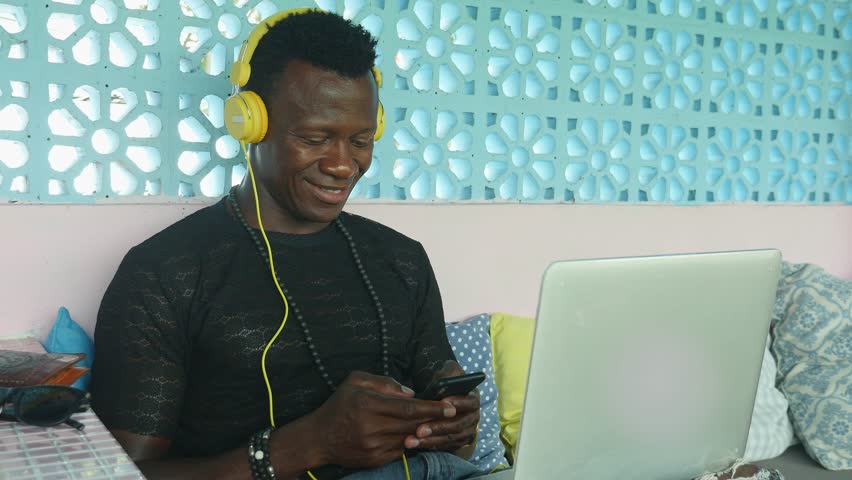 Young happy and attractive black African American hipster man networking relaxed with mobile phone and laptop computer smiling in digital nomad job success and internet work lifestyle | Shutterstock HD Video #1027277978