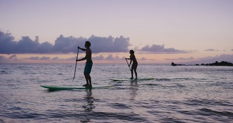 Two stand up paddlers enjoy tropical island sunset in pacific ocean, active couple stand up paddling together, sunset silhouettes