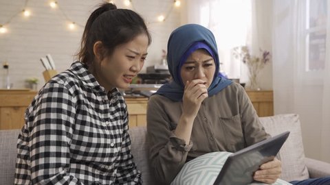 nervous young girl friends scary movie talking discussing. multi friendship leisure time relaxing at home together in kitchen sitting on sofa. asian lady muslim woman watching horror movie tablet