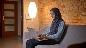Closeup shoot of young attractive muslim female teenager in hijab using the laptop and having a video call while sitting on the couch indoors at cozy home