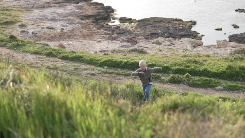 A blond boy walking by the sea. he explored the rocks and now climbs the top of the mountain. He walks through tall grass, beautiful light on video