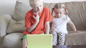 Children are looking at the tablet screen. They sit at home on the couch, in front of them on the table is an ipad in a green case. The boy and the girl do not take their eyes off the manitor.