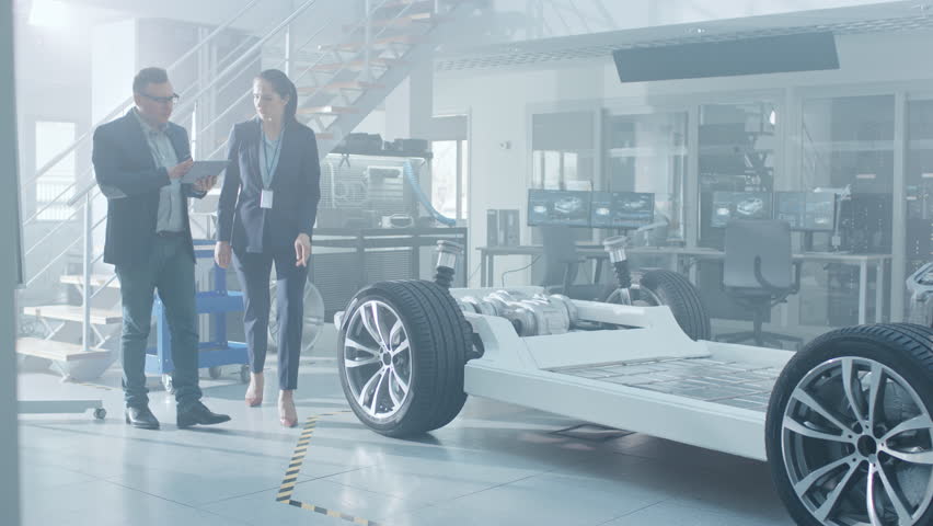 Female and Male Engineer Walk in a High Tech Development Facility Holding a Tablet Computer. They Pass an Electric Car Chassis Prototype with Batteries and Wheels.