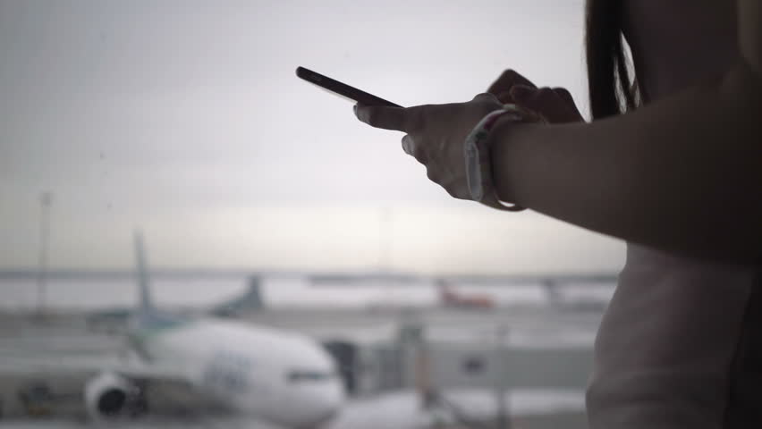 Woman using mobile phone at airport terminal and waiting for boarding the plane. UHD footage Royalty-Free Stock Footage #1027289939