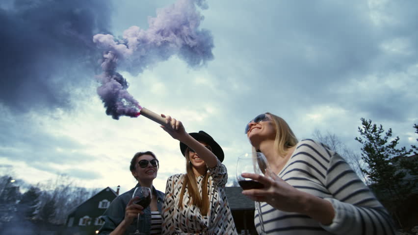 Low-angle medium shot of three young millennial chicks hanging out in twilight at suburban house party, enjoying red wine and waving smoke flare