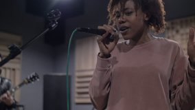 Lovely and young black woman is singing rock songs towards wired microphone while dancing to the music with energy and passion in recording studio.