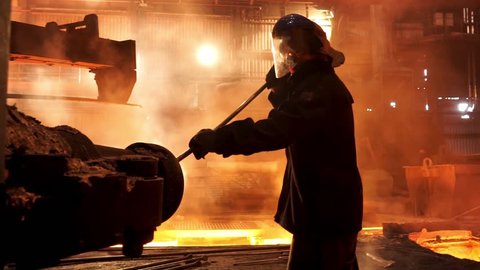 Steel worker removing slag from the electric induction crucible melting furnace at the metallurgical plant, hard work concept. Stock footage. Man in protective mask and uniform working with a poker.