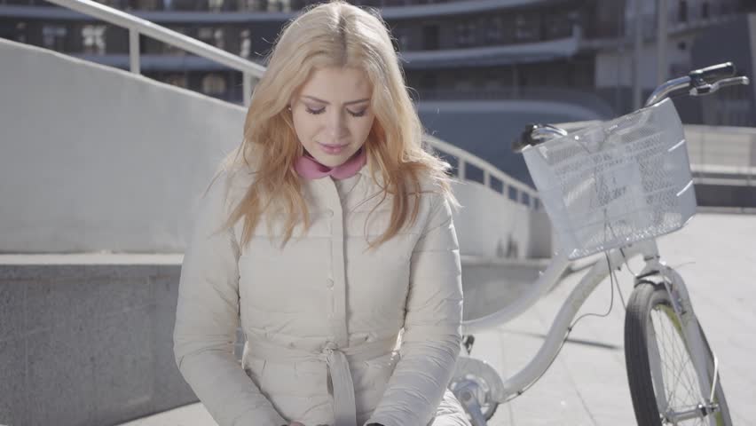 Portrait of a young beautiful girl with blond hair and in a light jacket sitting on a bench next to the bike. The girl is typing on a cell in good weather outdoors.