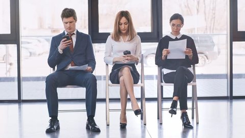 handsome man in formal wear using smartphone near attractive women talking while sitting on chairs with resumes