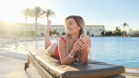 Attractive blonde woman in red swimsuit shaking head, listening music from her smartphone, while lying on deck chair in hotel swimming pool side area. Girl sunbathing and relaxing at resort.
