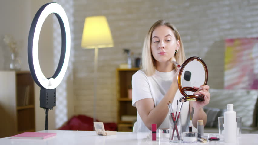 Lockdown of young Caucasian woman putting face powder with brush on her face while looking in mirror