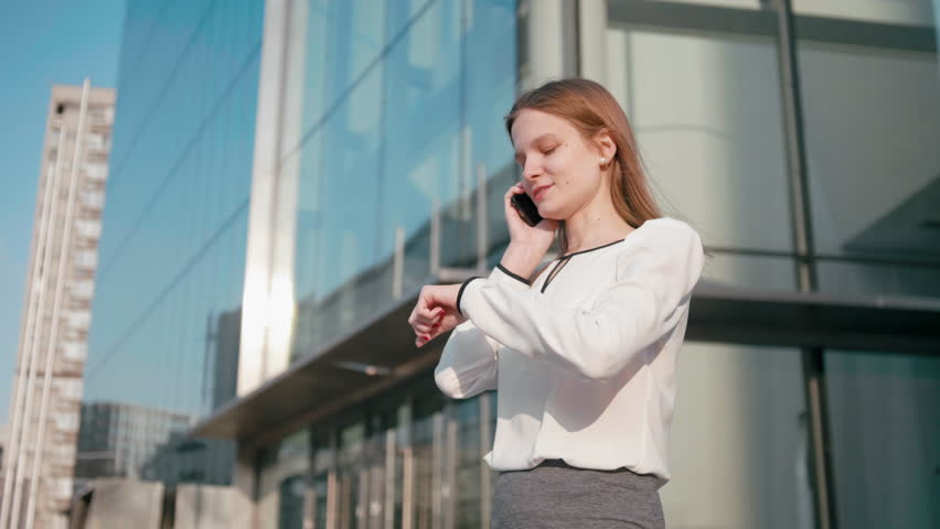 Adult Caucasian Confident Young Business Woman in White Shirt is Talking on Phone Outside near Modern Office Building. Medium Long Low Angle 4K Slow Motion Corporate Shot with Moving Around 360 Camera