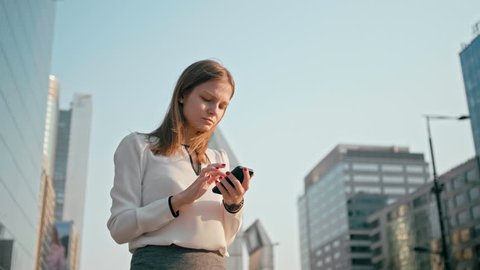 Adult Caucasian Confident Young Business Woman is Using Smartphone App Outside near Modern Office Building. Medium Low Angle 4K Slow Motion Corporate Shot with Moving Camera Around