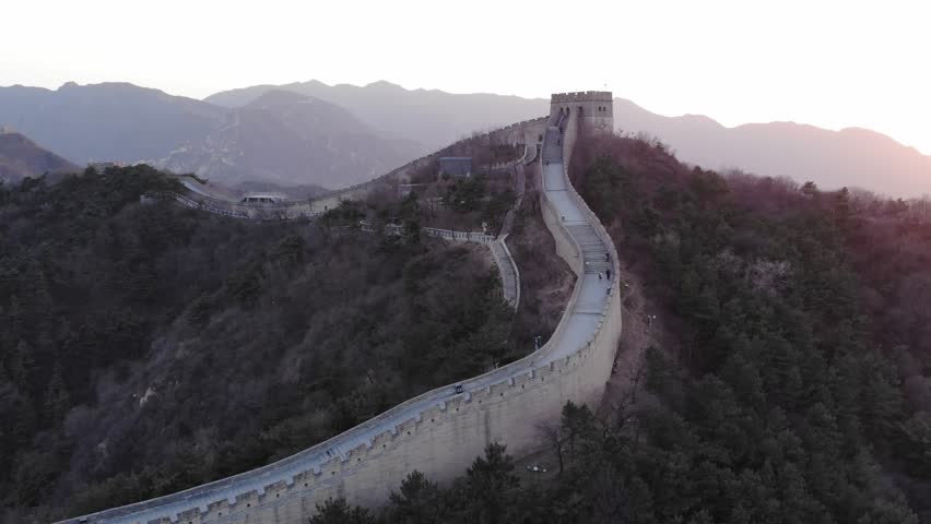 Aerial shot of Great Wall of China at sunset, tower seen on top of hill. Few tourists climb steps up to building at evening hour, dim sun shine on background, camera fly around