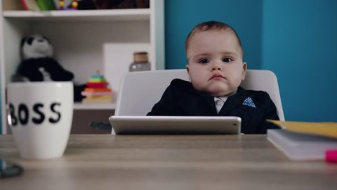 close up view of lovely toddler boy in business suit with extremely cute cheeks sits at the table with tablet by the baby boss?s mug. Successful baby, childhood happiness, little wonder. Child?s