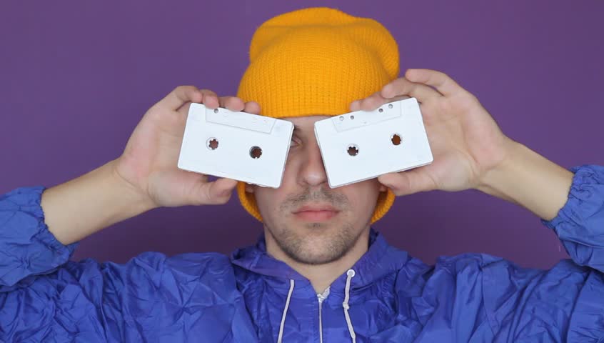 young man in a yellow cap and blue jacket holds vintage audio cassettes near his eyes