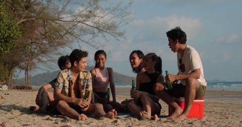 Asian teen group drinking beverage and sitting seaside together at beach summer with sunset background. Young asia happy emotion and anniversary celebration. 4K resolution and slow motion.