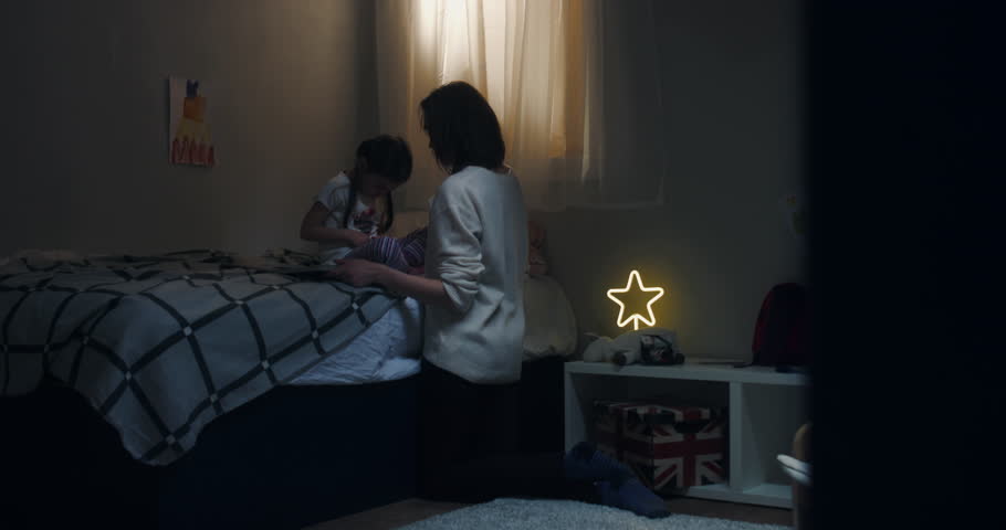 DOLLY IN Portrait of cute little girl sits on bed, reading a book together with her mother at home, evening shot, bedroom interior. 4K UHD RAW FOOTAGE