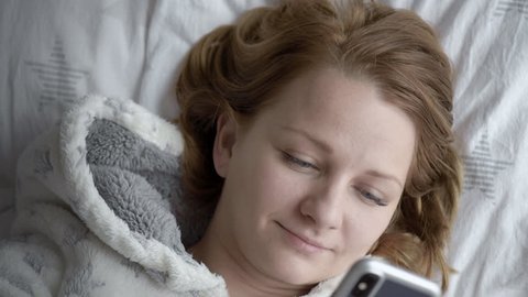 Happy Woman are using Mobile Phone on bed in morning. Girl in pajamas checking social apps with Smartphone. Smiling woman surfing net with Cellphone at home - top view.