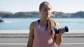 Sporty young woman exercising with dumbbells on riverside. Front view of beautiful blonde training outdoor. Concept of sport