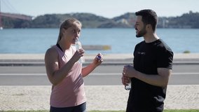 Sporty young people drinking water from plastic bottles. Tired young bearded man and blonde woman resting after training on embankment. Healthy lifestyle concept