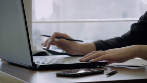 Close-up of the female hands of the secretary, sits in the office at the desk, typing on a laptop, making notes in pencil on a sheet of paper. Slow motion, unrecognizable person