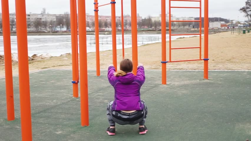 Athlete squats with dumbbells. Beautiful girl is engaged in sports. A young woman trains on the street. A woman with a sporty body squats. Workout. Playground on the street.
