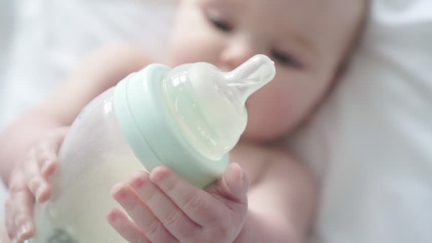 Pretty baby girl drinks milk from bottle lying on bed. Child weared diaper in nursery room. | Shutterstock HD Video #1027391027