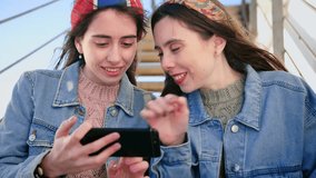 Two engaging young Hispanic girls watch new video on YouTube application, liked it and shared, using new smart phone device, city landscape on the background