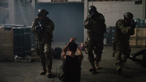 Three special ops military SWAT team members appear from dark smoke walking towards camera shooting and killing bad guys in a fight in a warehouse under dramatic daytime lighting. Shot on RED camera