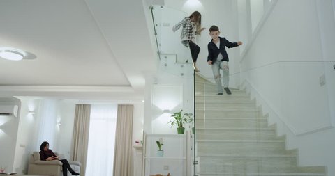 In a modern house with a large living and white stairs two kids running down to the first floor in the morning to their mother and small sister , hugging each other they start a good day.