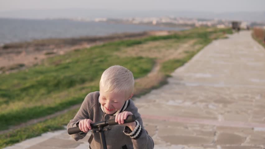Funny boy blond rides a scooter along the promenade. A child rolls on a scooter on the road by the sea. Spring walk by the sea family