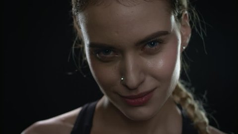 Sport woman portrait on black background. Close up of sweat woman face looking camera in dark gym. Exhausted fitness model wiping sweat from face in studio. Closeup of tired fit girl face posing