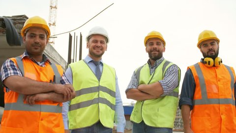 Tracking shot of group of confident construction site workers in safety vests and hard hats walking towards camera and posing in front of unfinished concrete building