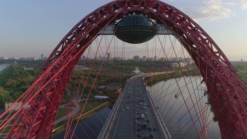Aerial photography/ drone shooting: cable-Stayed car bridge with a red metal arch over the river at sunset, heavy traffic, the camera flies under the arch over the highway. Picturesque Bridge